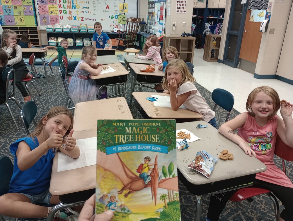 students sitting at desks smiling and giving thumbs up while the teacher holds up a Magic Treehouse book that she is reading to them